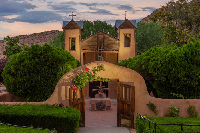 A historic adobe shrine with two wooden bell towers seen in a walled adobe courtyard with ornate rose-covered wooden doors and trees throughout.