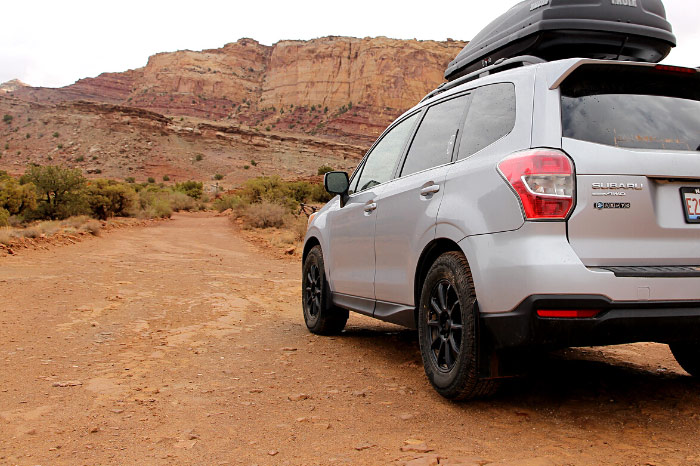 A car on remote graded dirt road with views of canyons and rock formations ahead.