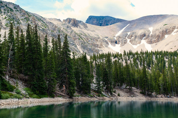 An emerald-colored alpine lake with pine trees surrounding it, sitting at the base of a massive mountain with patches of snow throughout.