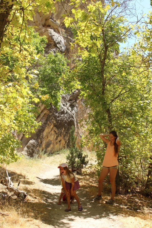 Mom and two young daughters resting in the shade along the narrow, sandy Box Canyon Trail, surrounded by trees, shrubs, and a canyon in the background.