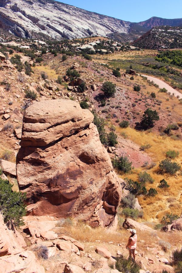 View of Cub Creek Road in Dinosaur National Monument, Utah, with colorful rocky hills in the distance. Photo taken from a higher vantage point at the the Cub Creek Petroglyphs site, with rocks and boulders in the foreground and a small figure standing below, taking in the views and scanning the cliff faces for more petroglyphs.