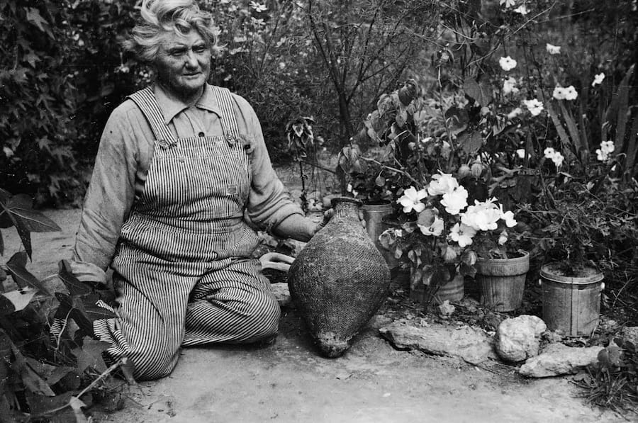 Josie Morris in overalls, sitting on the ground outside, surrounded by flowers, holding a vase at her Cub Creek homestead.