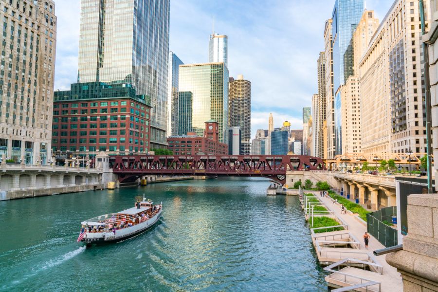 Scenic view of the Chicago Riverwalk with city skyline, bridge, and passing boat.