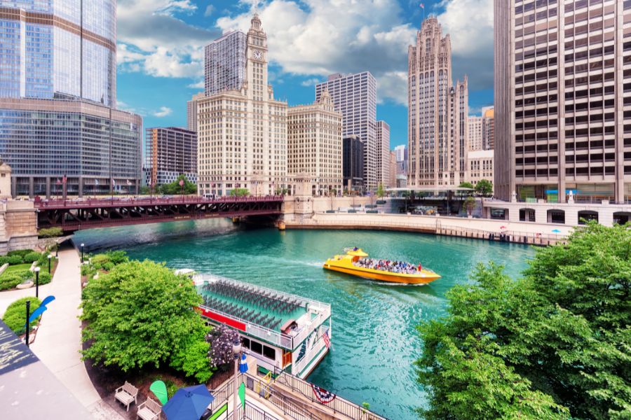 View of a boat cruising on the Chicago River along the Chicago Riverwalk, with downtown skyscrapers in the background and trees in view.