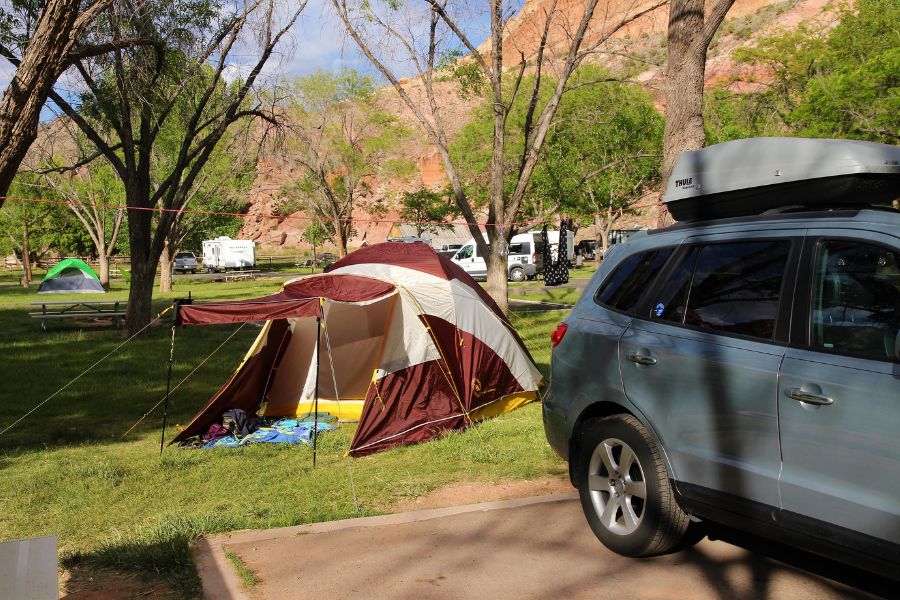 Car parked beside a tent at Fruita Campground in Capitol Reef National Park, set on a grassy field with trees and red rock cliffs in the distance.