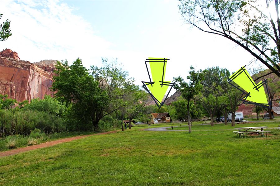 View of the grassy field at Fruita Campground in Capitol Reef National Park with red rock cliffs in the background and arrows pointing to the Gifford House and Fruita Barn in the distance.