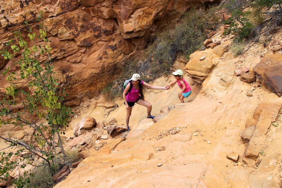 Mom reaching out to help her younger daughter descend a steep, sandy, slippery section of the Tanks spur trail, surrounded by desert vegetation, navigating the tricky terrain together.