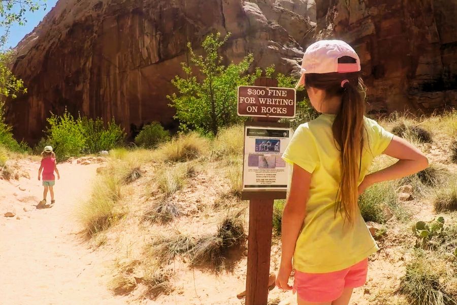 An older child pauses to read an informational sign near the Capitol Gorge trailhead as a younger sibling continues up the sandy wash, framed by a towering canyon wall on the right.