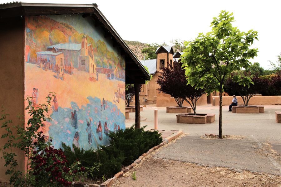 Part of the courtyard at El Santuario de Chimayó, featuring a large adobe wall adorned with a colorful, bright mural.