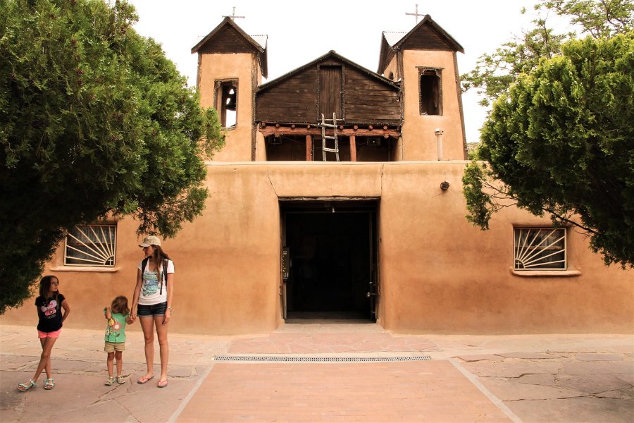 A mother and her children standing in the courtyard in front of the main adobe chapel at El Santuario de Chimayó, with the chapel’s open doorway, wooden roof accents, and a ladder leading to the roof visible.