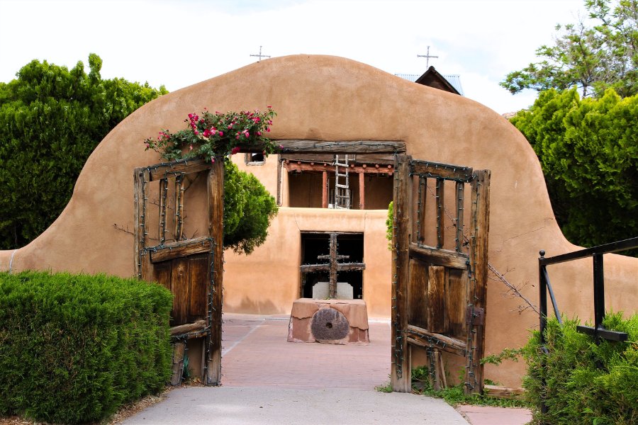 Entrance into a walled courtyard at El Santuario de Chimayo through an arched adobe gate with rustic wooden doors and roses growing around them.