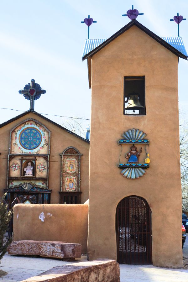 The Santo Niño de Atocha Chapel at El Santuario de Chimayó, showing richly decorated adobe buildings with arched metal doors, painted details, and a bell above.