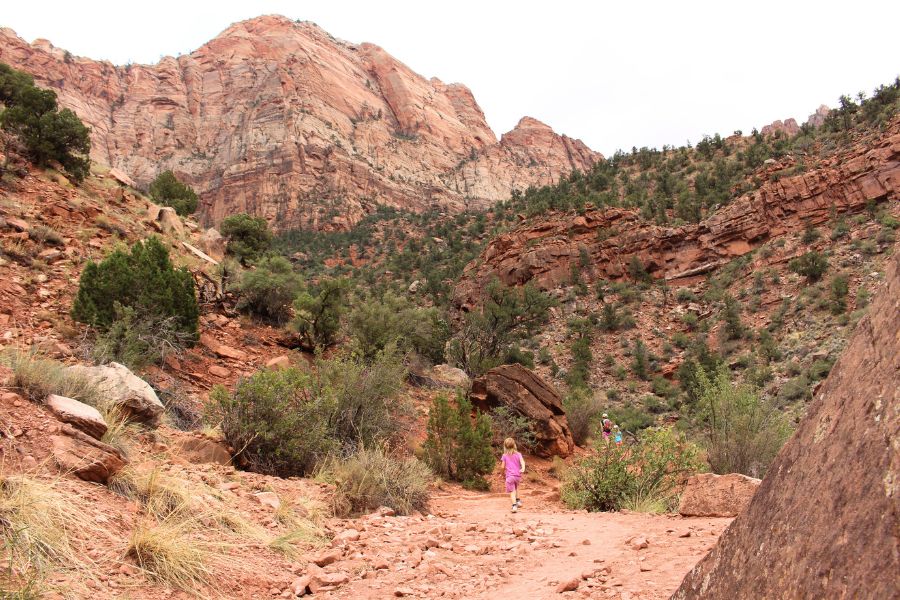 Mom and kids in the distance, hiking uphill toward the plateau, surrounded by dry desert landscape and canyon cliffs.