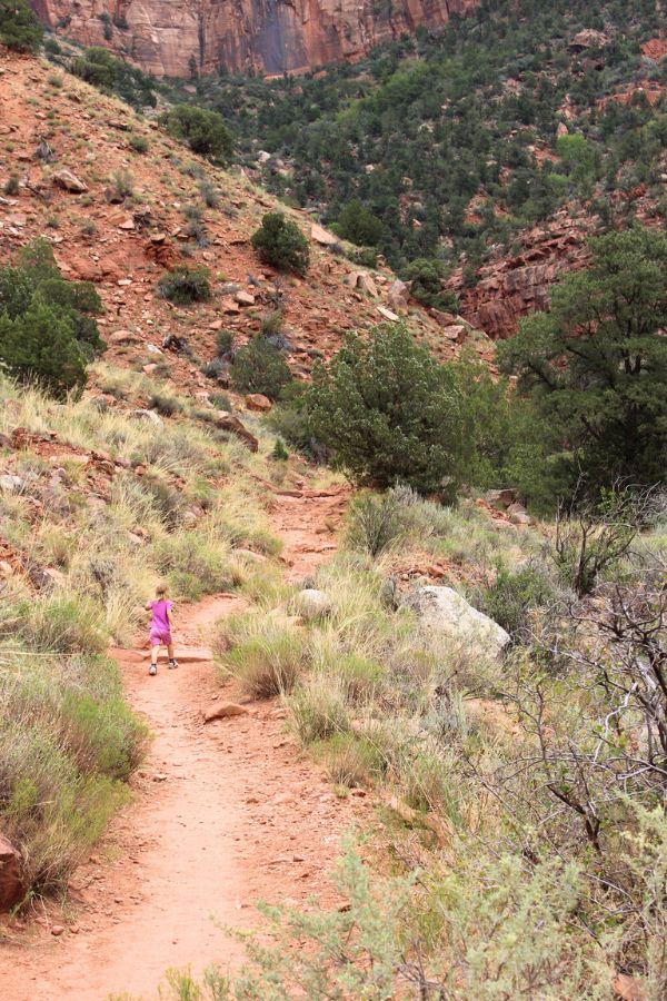 Daughter hiking along a narrow section of trail with hills, cliffs, and desert plants ahead.