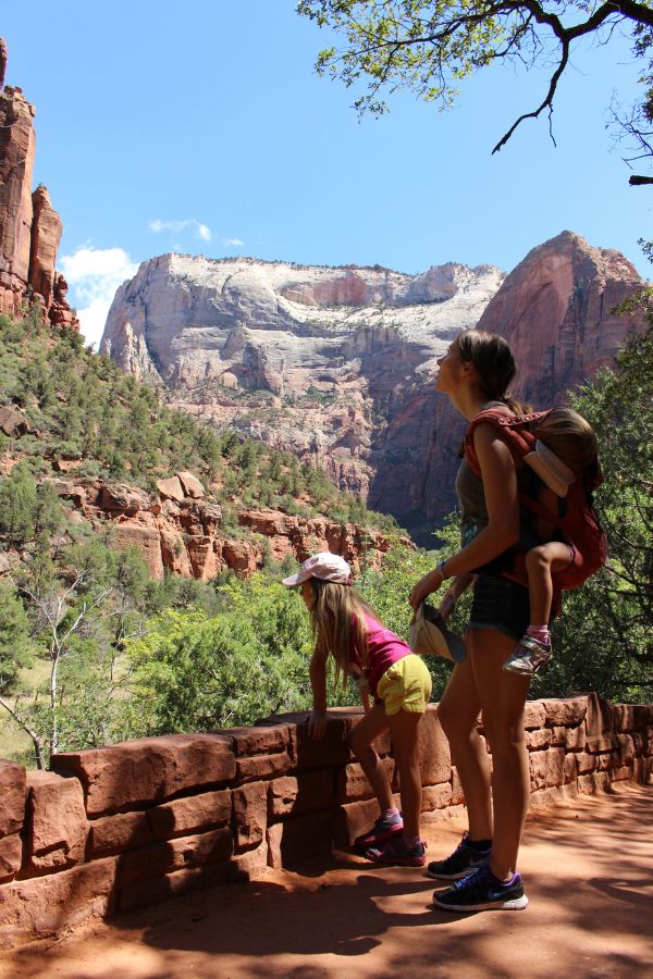 Mother carrying a child in a backpack carrier while standing with another daughter on the trail to natural pools in Zion National Park, looking toward towering sandstone cliffs.