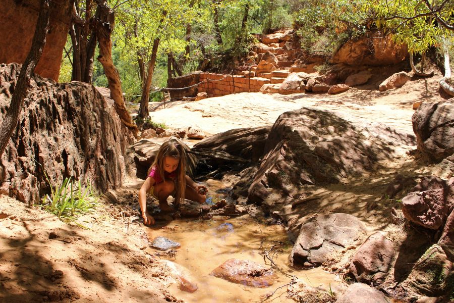Child exploring a shallow stream near the chained overlook at Middle Emerald Pools with slickrock and trees surrounding the area.