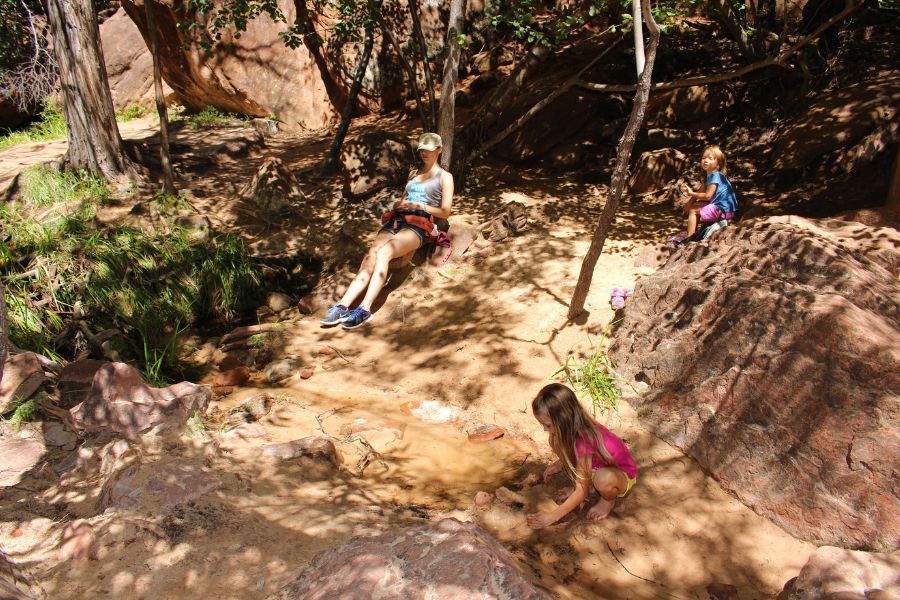 Family resting in a shaded sandy area near Middle Emerald Pools with rocks and vegetation surrounding them.