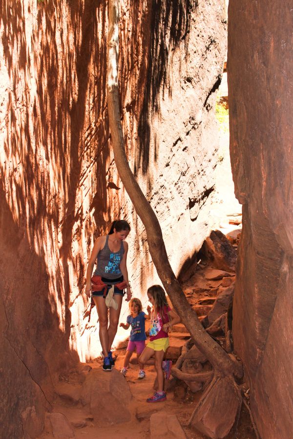 Family passing through a narrow rock opening on the Middle Emerald Pools trail surrounded by sand and shaded cliffs.