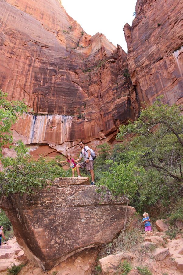 Hikers standing on a large boulder along the Upper Emerald Pool Trail beneath towering cliffs, and another family member standing to the right of the massive boulder.