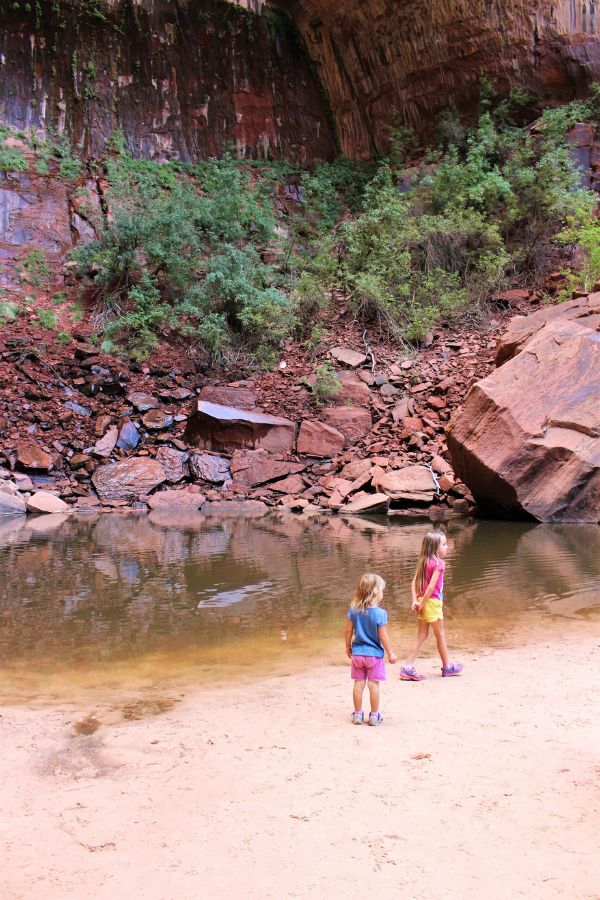 Children standing near the shoreline of Upper Emerald Pool with boulders around and cliffs rising behind them.