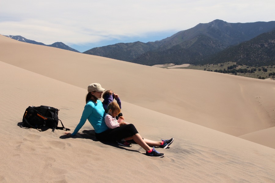 Family resting on a sandy ridge in Great Sand Dunes National Park backed by rugged mountain peaks.