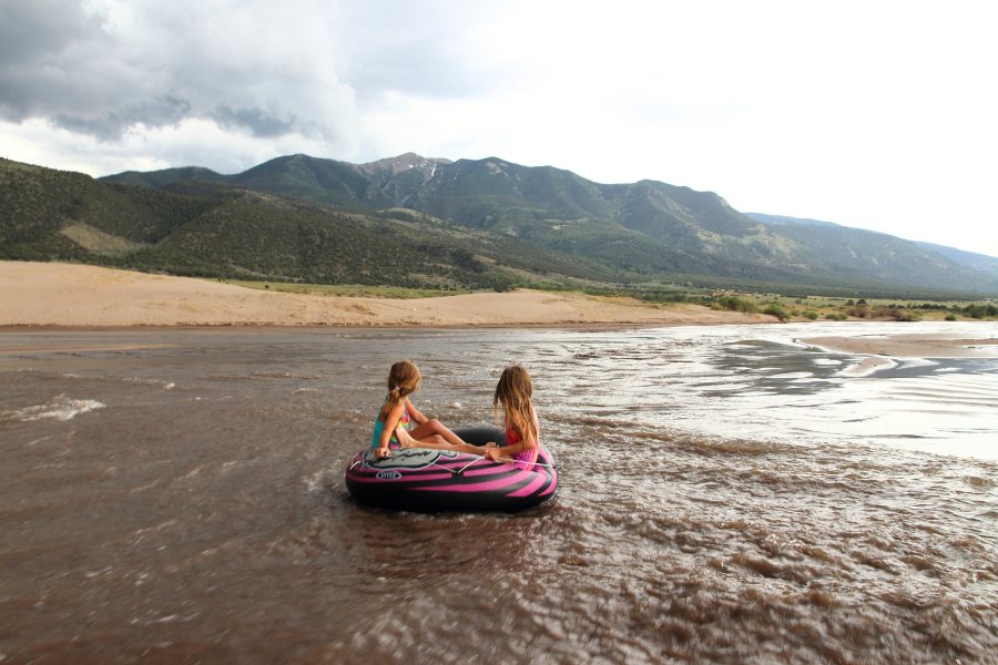 Two kids floating on an inflatable tube down the wide and shallow Medano Creek, with sandy shore and forested hills in view across from the stream.