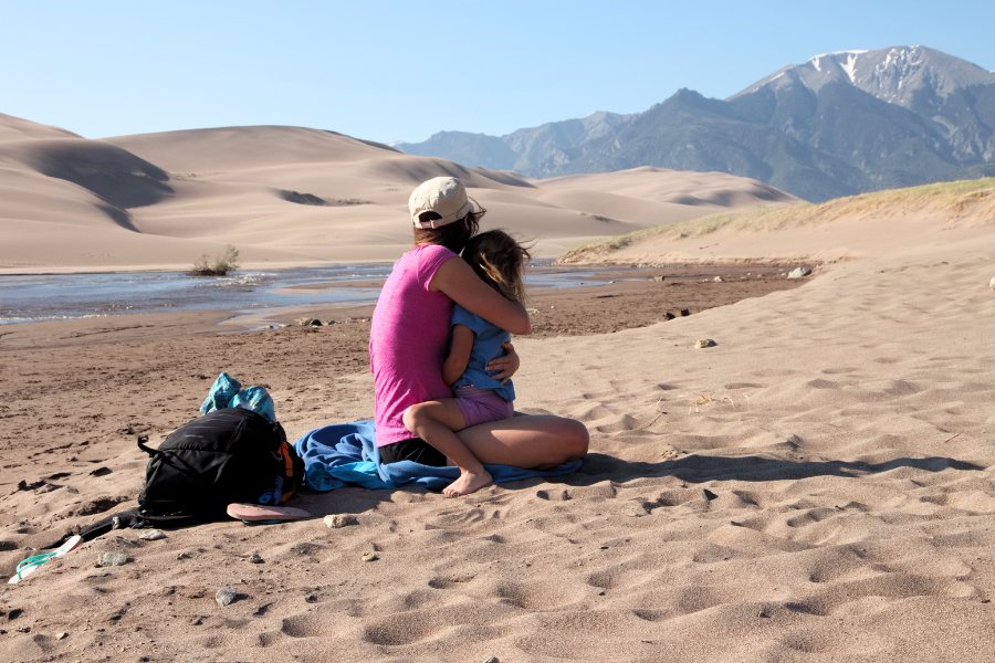 Mom holding a toddler on the sand next to Medano Creek, with a massive dunefield rising across the stream, and snow-capped mountains in the background up ahead.