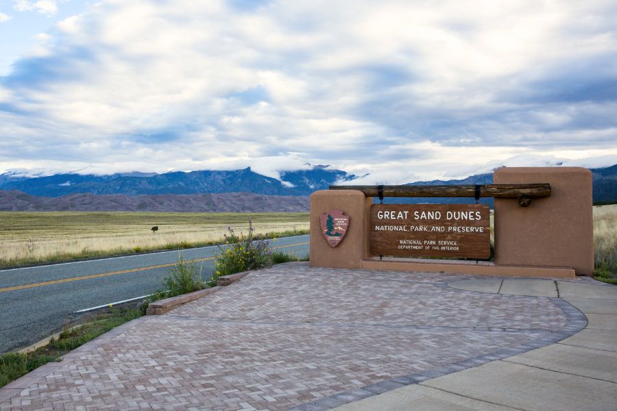 The entrance and welcome point to Great Sand Dunes National Park and Preserve in Colorado, showing the sign for the park, distant towering sand dunes with tall mountains behind them, and a green valley and a small section of road leading up to the distant dunefield.