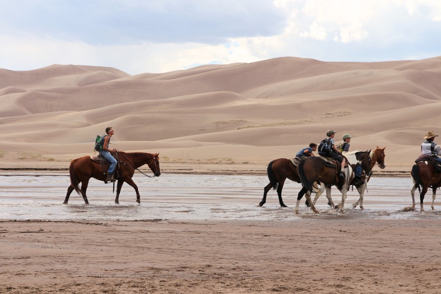 Horseback riders moving through a wide shallow creek in Great Sand Dunes National Park, with massive sand dunes rising just behind them.