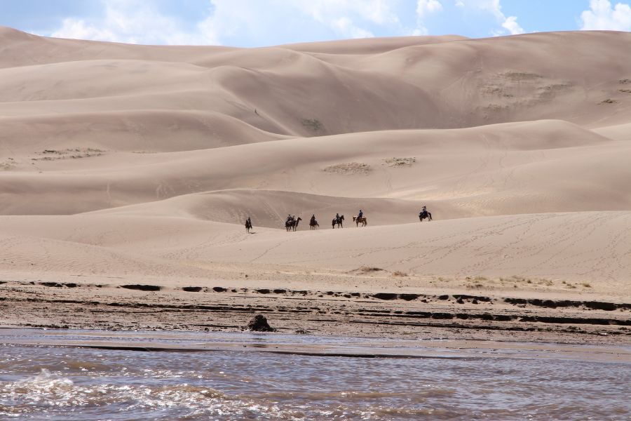 Horseback riders moving through the lower part of the massive dunefild in Great Sand Dunes National Park, with flue sky and Medano Creek flowing at the dune base.