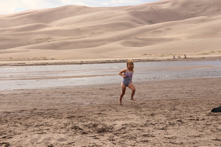 Child running in the sand next to Medano Creek, with massive dunes rising in the background across the water.