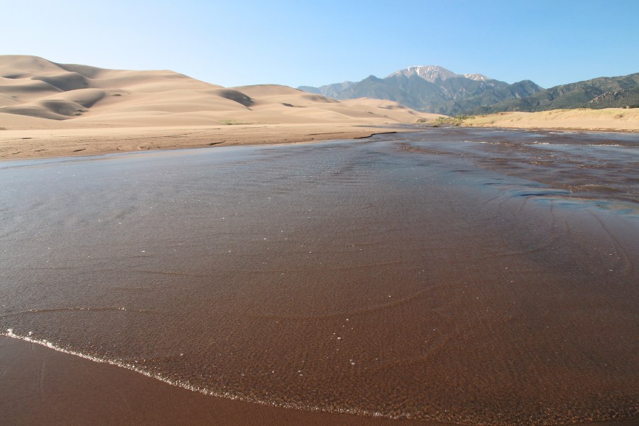 Shallow, slow-flowing Medano Creek, with dunes rising on the left side across the water, and snow-capped mountains in the background up ahead.