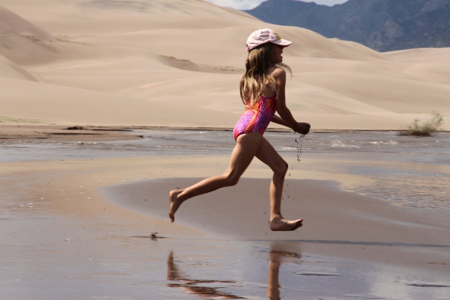 Child running joyfully through Medano Creek in Great Sand Dunes National Park, with dunes and mountains in the background.