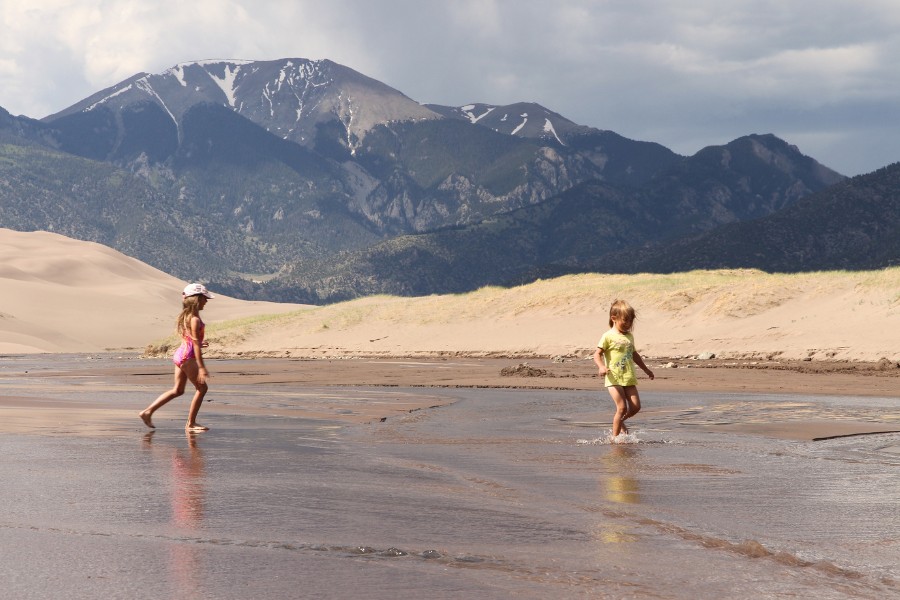 Kids running through the shallow, gently flowing Medano Creek, with sandy shores all around, dunes rising to the left, and snow-capped mountains rising in the background up ahead.