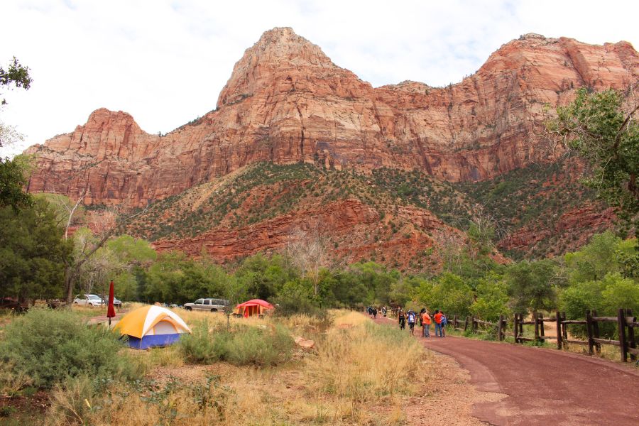 The beginning of the paved Pa'rus Trail in Zion National Park near the Visitor Center, showing a wide paved path, tents pitched along the left side, lush vegetation all around, and red rock cliffs rising ahead.