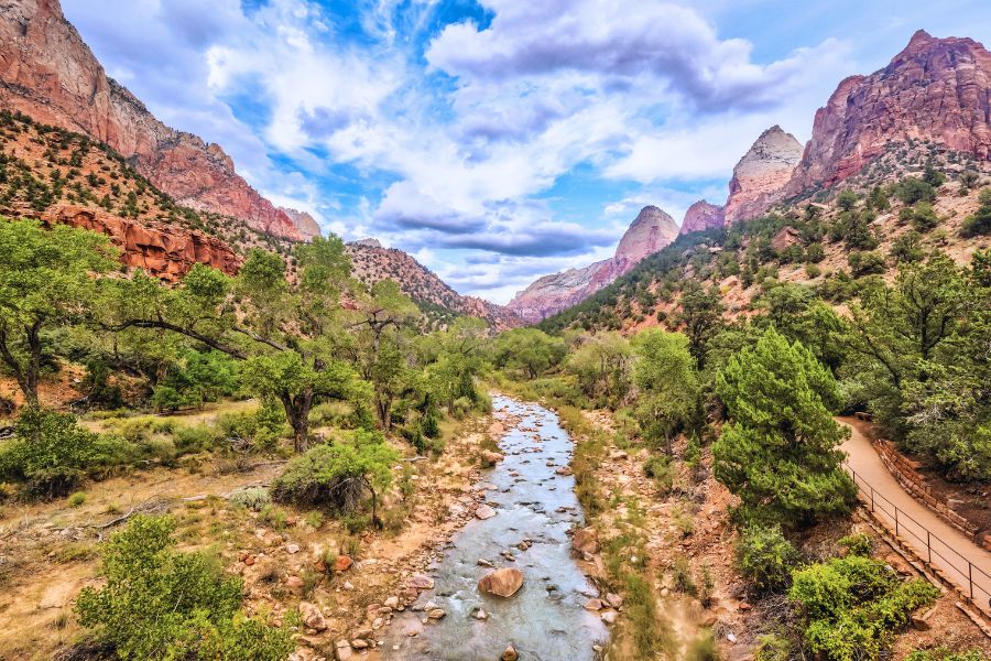 A paved path along the Virgin River in Zion Canyon with lush vegetation along both riverbanks and sculpted cliffs rising on both sides.