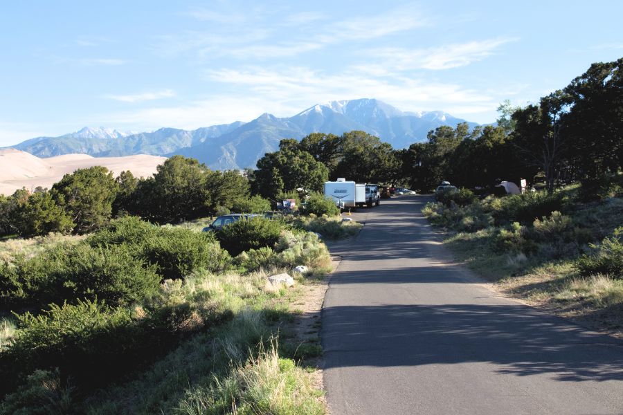 Road through the Pinon Flats Campground in Great Sand Dunes National Park, with lots of vegetation around, and dunes and mountains in the background.