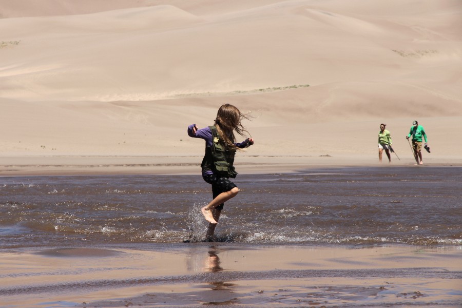 Child running joyfully through Medano Creek, with the dunefield rising just behind across the water, and visitors walking on the sand on the other side of the stream.