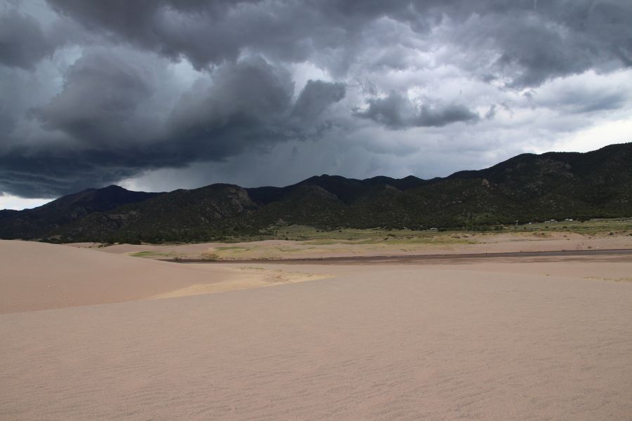 Sand dunes stretching across the valley floor with mountains behind and dark stormy clouds above.
