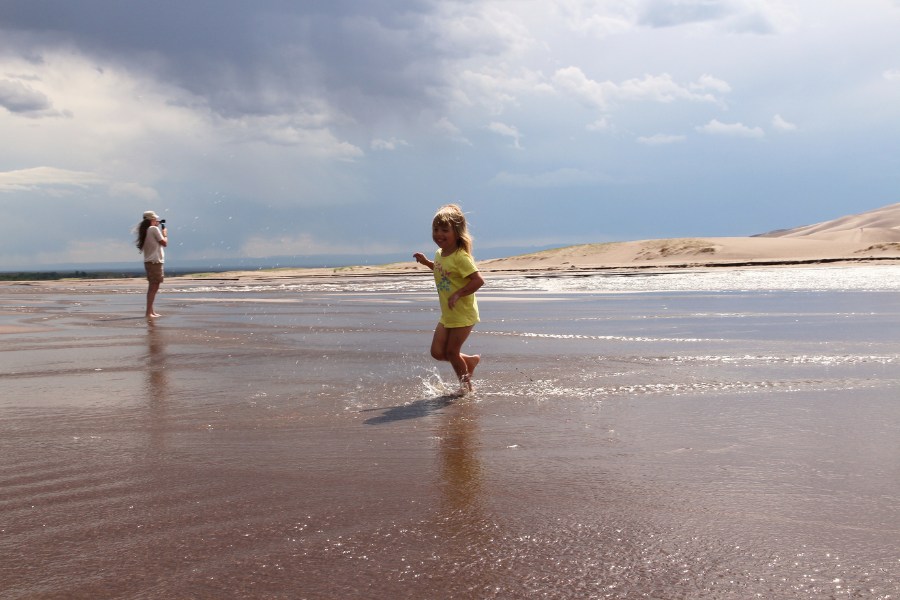 Mom and daughter standing in the middle of the shallow and gentle flowing seasonal Medano Creek, with storm clouds building up above, and dunes beginning to rise across the water.
