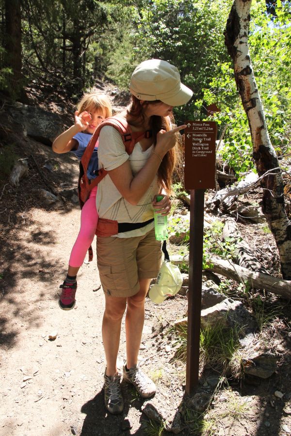 Hiker carrying a child at a shaded trail junction with directional sign for the Mosca Pass, Montville, and Wellington Ditch trails in Great Sand Dunes National Park.