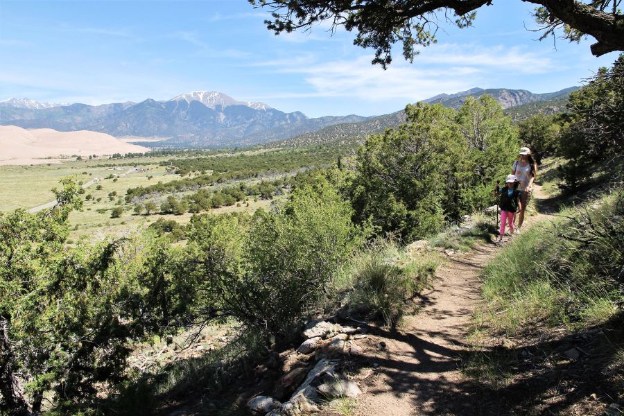 Hikers on narrow Wellington Ditch Trail with dunes and mountains in view.