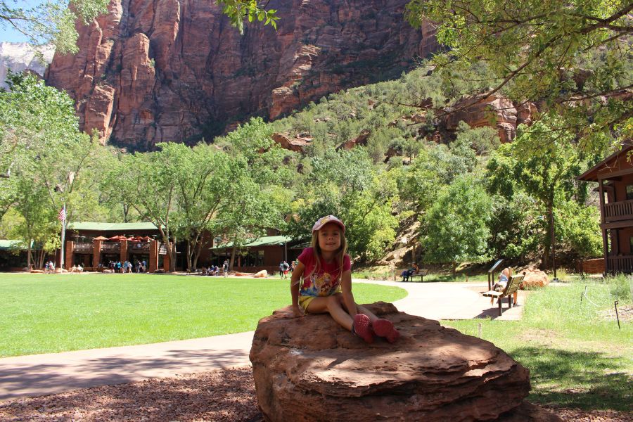 Child sitting on a small boulder at the Zion Lodge lawn area in Zion National Park, with the lodge visible across the large lawn, and Zion Canyon cliffs rising in the background.