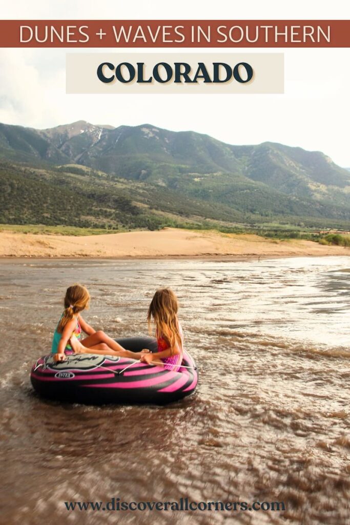 Two kids floating on an inflatable tube down the wide and shallow Medano Creek in Great Sand Dunes National Park, with sandy shore and forested hills in view across from the stream. Text overlay - Dunes + waves in southern Colorado.